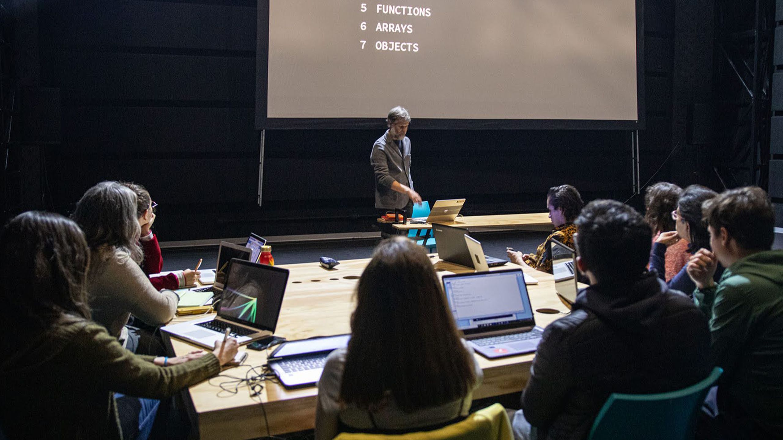 Casey Reas teaching a programming class, students at laptops around a long table below a projected slide