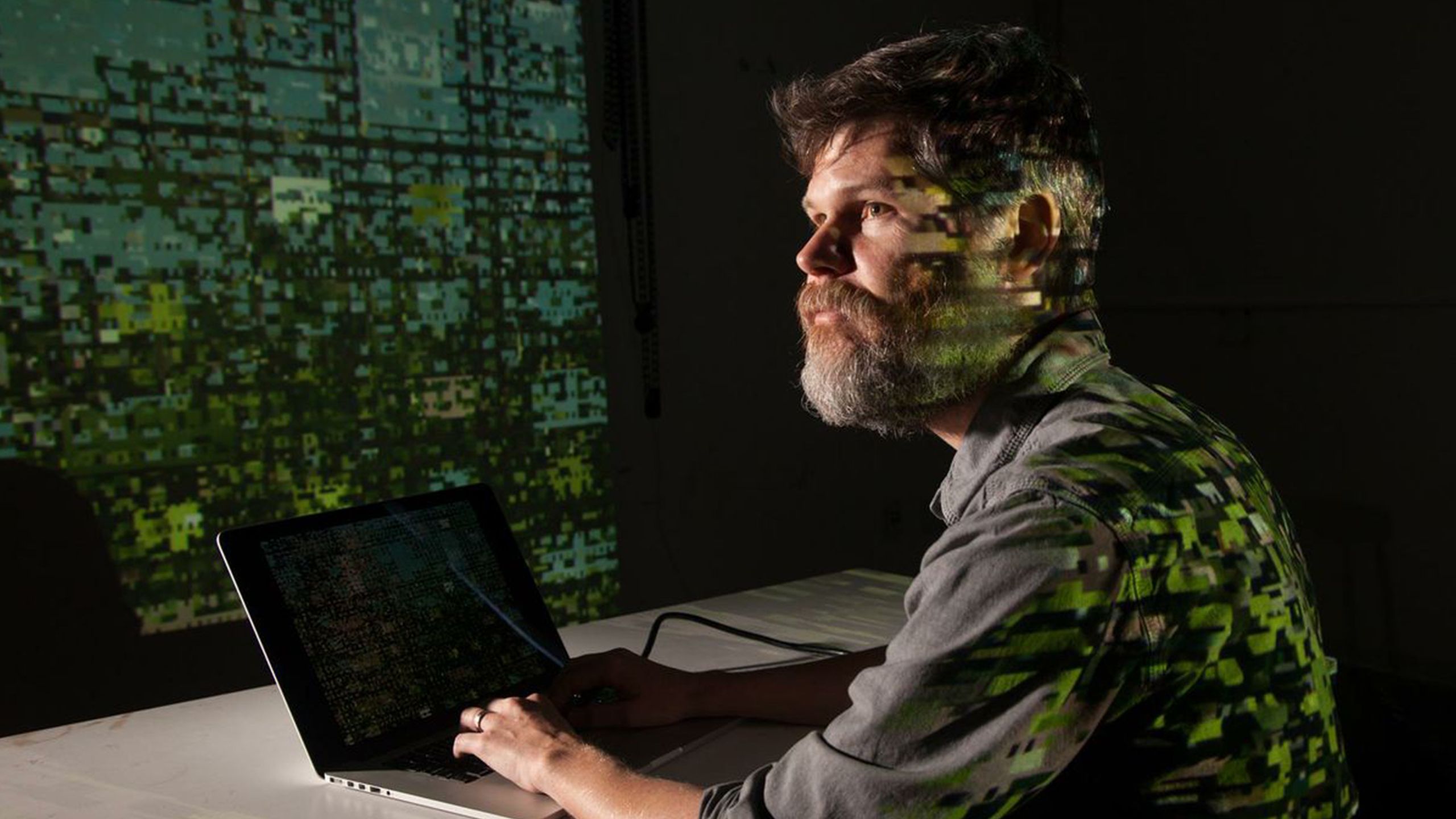 Casey Reas at a laptop, a projected grid of green and black pixels falling across his face